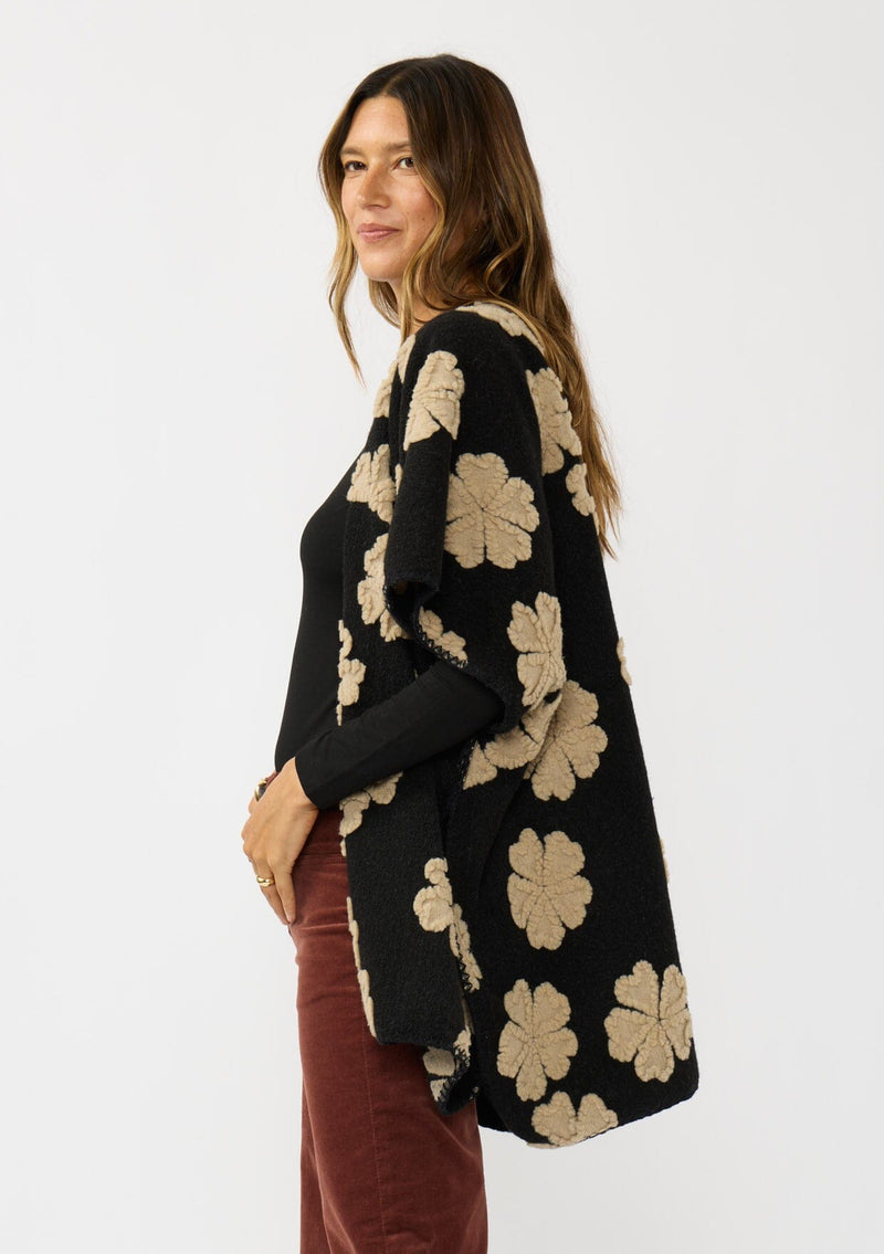 [Color: Black/Taupe] A side facing image of a brunette model wearing a fuzzy floral poncho with sleeve openings and an open front. A unique and casual throw on for the fall season. 