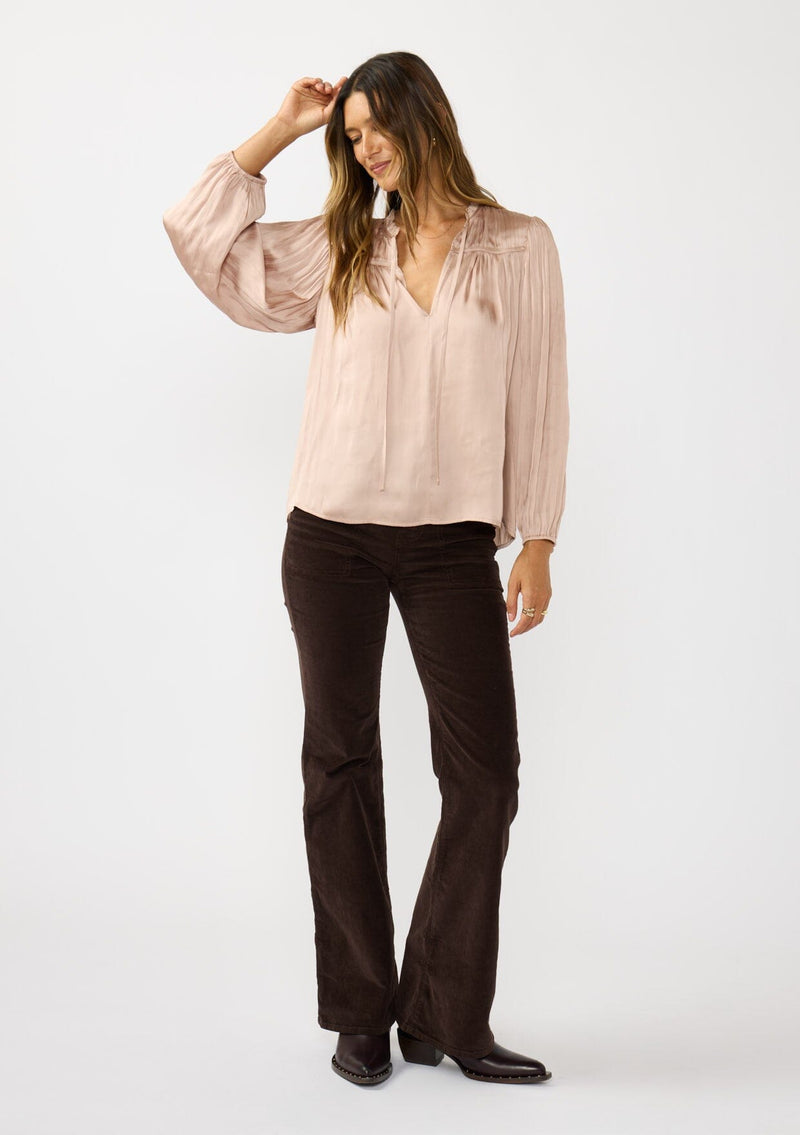 [Color: Champagne] A front facing image of a brunette model wearing silky rose gold blouse with a pleated yoke and sleeve. Featuring a relaxed split v-neckline for a laid back vibe. Effortless and charming for the Holiday season. 
