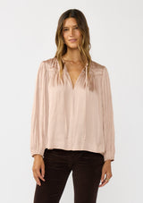 [Color: Champagne] A front facing image of a brunette model wearing silky rose gold blouse with a pleated yoke and sleeve. Featuring a relaxed split v-neckline for a laid back vibe. Effortless and charming for the Holiday season. 