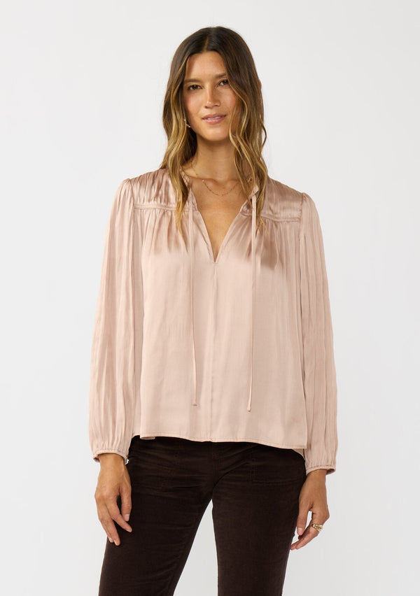 [Color: Champagne] A front facing image of a brunette model wearing silky rose gold blouse with a pleated yoke and sleeve. Featuring a relaxed split v-neckline for a laid back vibe. Effortless and charming for the Holiday season. 
