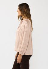 [Color: Champagne] A side facing image of a brunette model wearing silky rose gold blouse with a pleated yoke and sleeve. Featuring a relaxed split v-neckline for a laid back vibe. Effortless and charming for the Holiday season. 