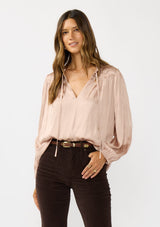 [Color: Champagne] A front facing image of a brunette model wearing silky rose gold blouse with a pleated yoke and sleeve. Featuring a relaxed split v-neckline for a laid back vibe. Effortless and charming for the Holiday season. 