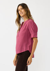 [Color: Berry] A side facing image of a brunette model wearing a pink puff sleeve top with gold metallic threads, a smocked detail on sleeve, and a button front. 