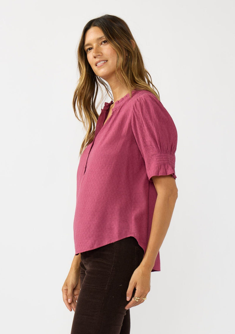 [Color: Berry] A side facing image of a brunette model wearing a pink puff sleeve top with gold metallic threads, a smocked detail on sleeve, and a button front. 