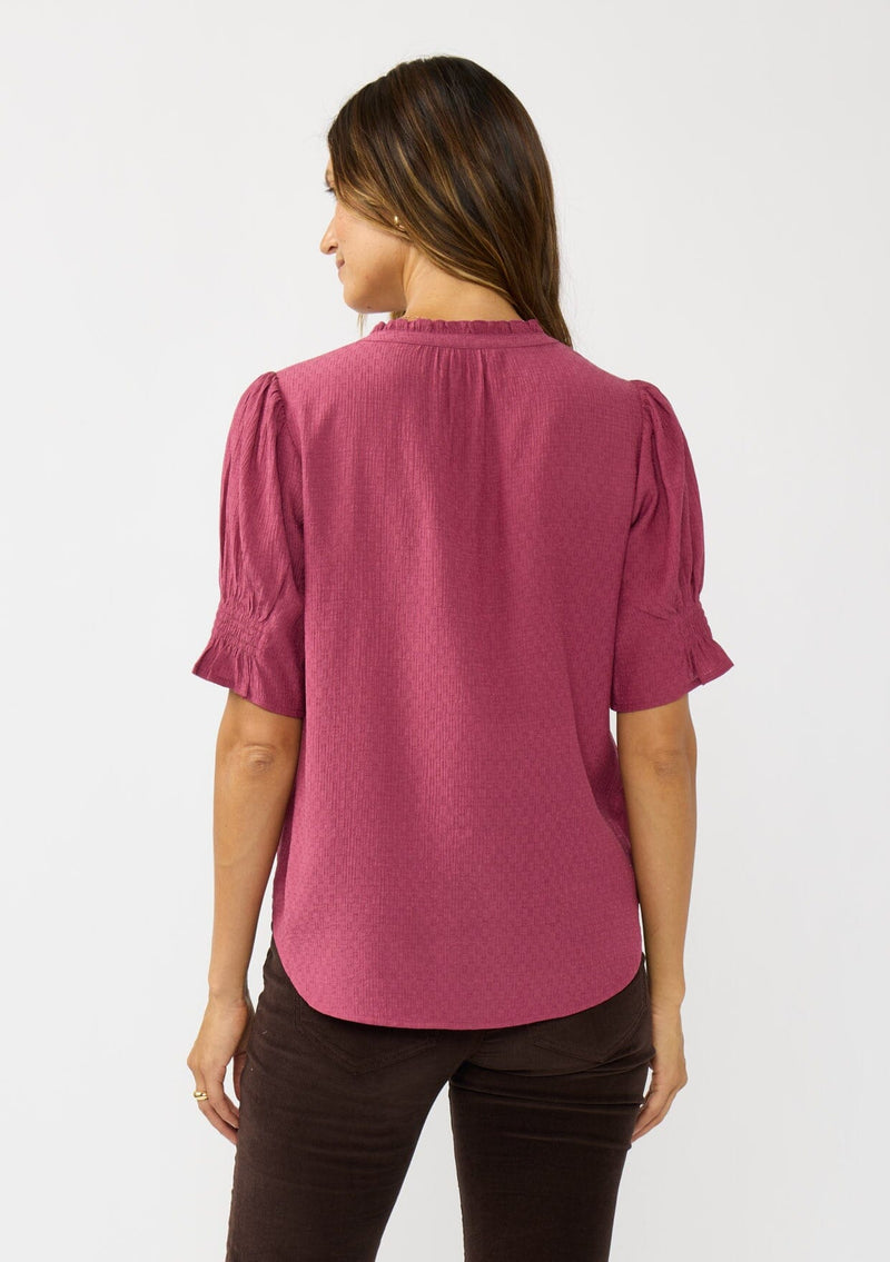 [Color: Berry] A back facing image of a brunette model wearing a pink puff sleeve top with gold metallic threads, a smocked detail on sleeve, and a button front. 