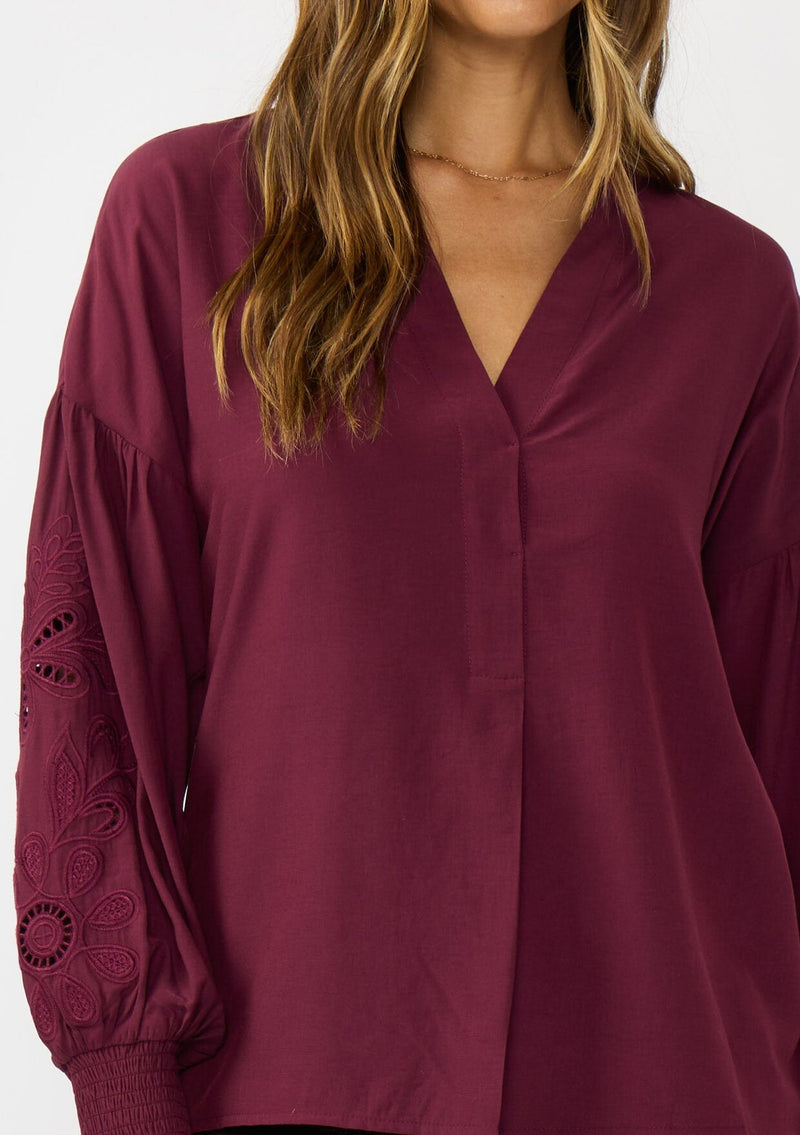 [Color: Merlot] A detail image of a brunette model wearing burgundy blouse with embroidery. Featuring long bishop sleeves, smocked cuffs, and a split v-neckline. An elevated boho blouse for the office or casual outings. 