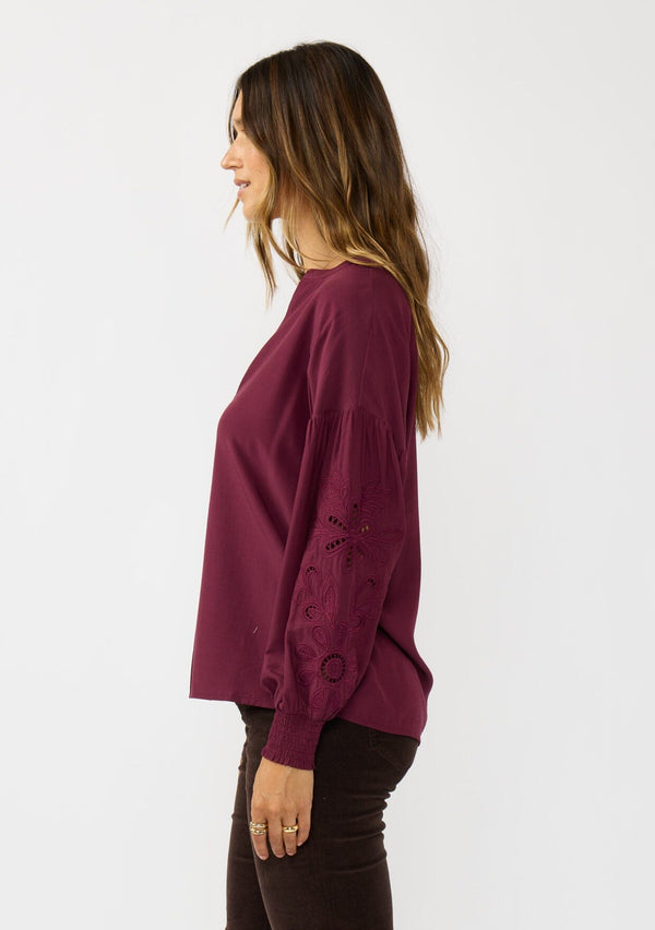 [Color: Merlot] A side facing image of a brunette model wearing burgundy blouse with embroidery. Featuring long bishop sleeves, smocked cuffs, and a split v-neckline. An elevated boho blouse for the office or casual outings. 