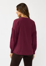 [Color: Merlot] A back facing image of a brunette model wearing burgundy blouse with embroidery. Featuring long bishop sleeves, smocked cuffs, and a split v-neckline. An elevated boho blouse for the office or casual outings. 