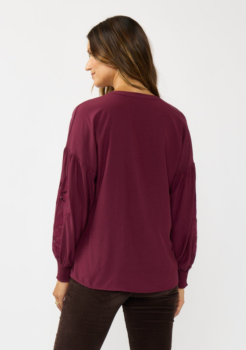 [Color: Merlot] A back facing image of a brunette model wearing burgundy blouse with embroidery. Featuring long bishop sleeves, smocked cuffs, and a split v-neckline. An elevated boho blouse for the office or casual outings. 