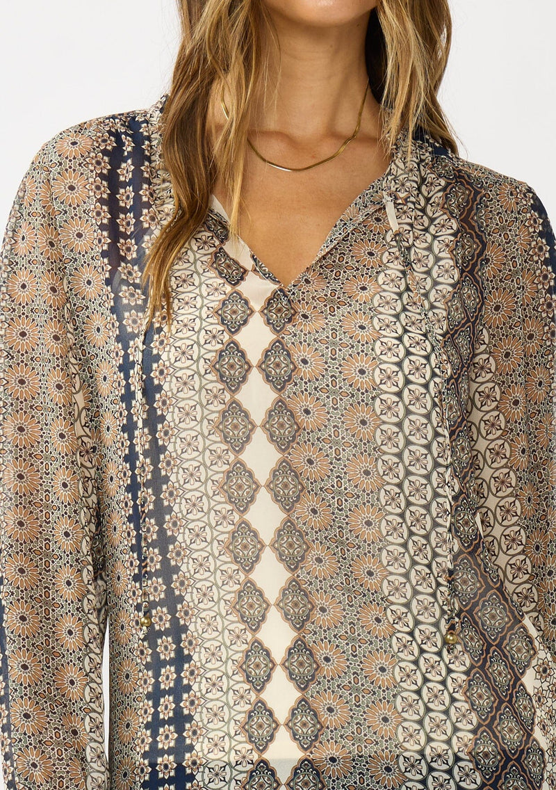 [Color: Natural/Navy] A detail image of a brunette model wearing a beige striped floral pattern blouse with a relaxed boho silhouette, split v-neckline with ties, and long sleeves with ruffled elastic cuffs. 