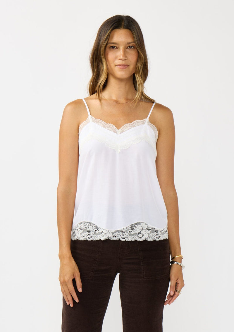 [Color: Chalk] A front facing image of a brunette model wearing a white cami with lace trims on the neck and hem, adjustable spaghetti straps, and a v-neckline. 