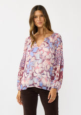 [Color: Natural/Dusty Blue] A front facing image of a brunette model wearing a pink and blue floral chiffon blouse with smocking on the neck and sleeve, and a split v-neckline with button closure. A standout flowy top for the fall season. 