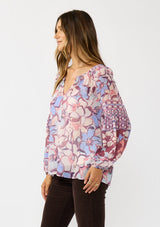[Color: Natural/Dusty Blue] A side facing image of a brunette model wearing a pink and blue floral chiffon blouse with smocking on the neck and sleeve, and a split v-neckline with button closure. A standout flowy top for the fall season. 
