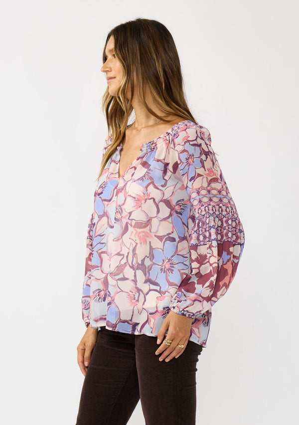 [Color: Natural/Dusty Blue] A side facing image of a brunette model wearing a pink and blue floral chiffon blouse with smocking on the neck and sleeve, and a split v-neckline with button closure. A standout flowy top for the fall season. 