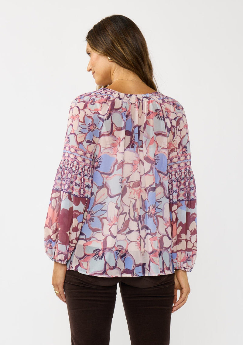 [Color: Natural/Dusty Blue] A back facing image of a brunette model wearing a pink and blue floral chiffon blouse with smocking on the neck and sleeve, and a split v-neckline with button closure. A standout flowy top for the fall season. 
