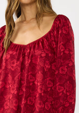 [Color: Red] A detail image of a brunette model wearing a holiday red floral jacquard blouse with a wide v-neckline, long sleeves, and an open back with tie detail. Elegant and sophisticated for holiday parties and special occasions. 
