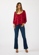 [Color: Red] A front facing image of a brunette model wearing a holiday red floral jacquard blouse with a wide v-neckline, long sleeves, and an open back with tie detail. Elegant and sophisticated for holiday parties and special occasions. 