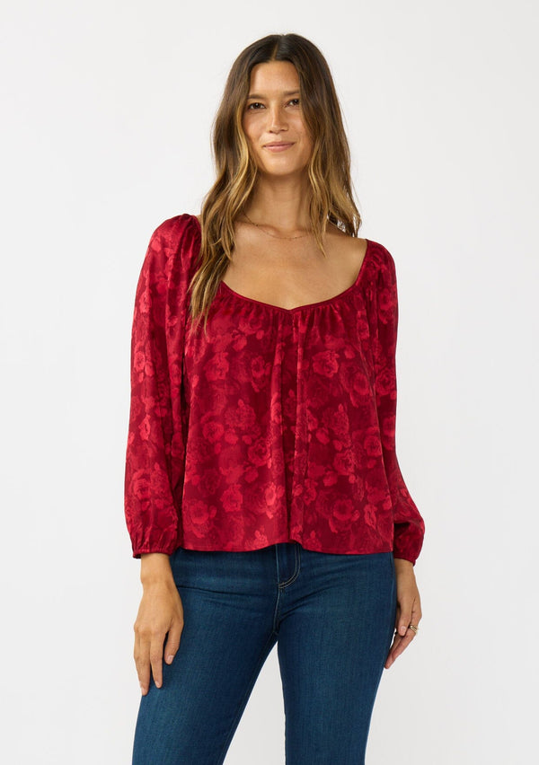 [Color: Red] A front facing image of a brunette model wearing a holiday red floral jacquard blouse with a wide v-neckline, long sleeves, and an open back with tie detail. Elegant and sophisticated for holiday parties and special occasions. 