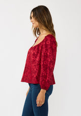 [Color: Red] A side facing image of a brunette model wearing a holiday red floral jacquard blouse with a wide v-neckline, long sleeves, and an open back with tie detail. Elegant and sophisticated for holiday parties and special occasions. 