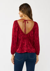 [Color: Red] A back facing image of a brunette model wearing a holiday red floral jacquard blouse with a wide v-neckline, long sleeves, and an open back with tie detail. Elegant and sophisticated for holiday parties and special occasions. 