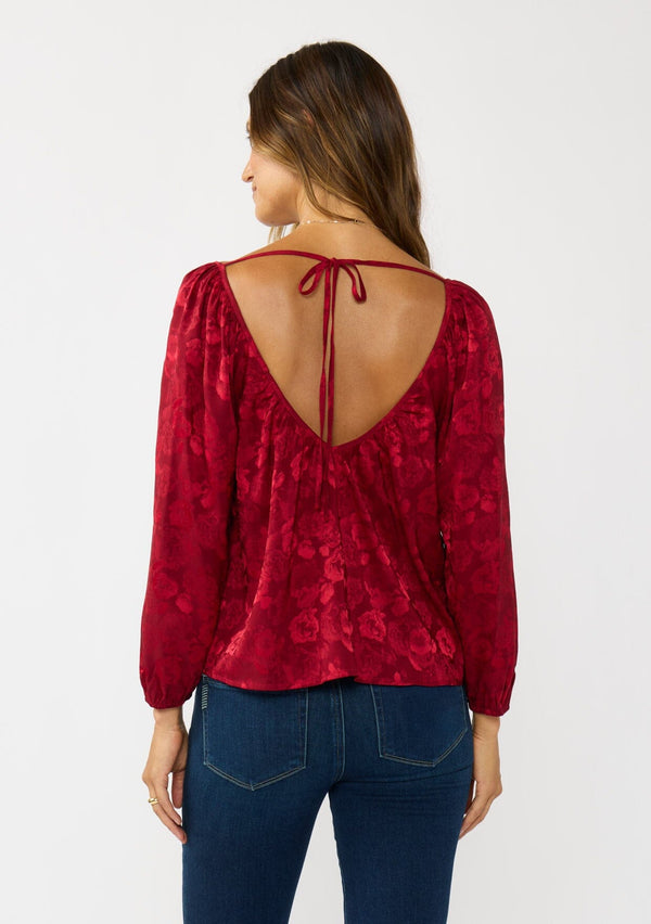 [Color: Red] A back facing image of a brunette model wearing a holiday red floral jacquard blouse with a wide v-neckline, long sleeves, and an open back with tie detail. Elegant and sophisticated for holiday parties and special occasions. 
