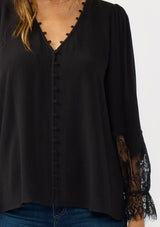 [Color: Black] A detail image of a brunette model wearing a solid black blouse with lace accented sleeves, button front, and v-neckline. A classic and versatile boho blouse with an elevated touch. 