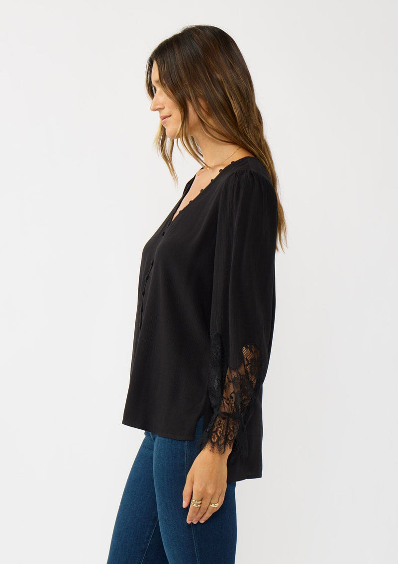 [Color: Black] A side facing image of a brunette model wearing a solid black blouse with lace accented sleeves, button front, and v-neckline. A classic and versatile boho blouse with an elevated touch. 