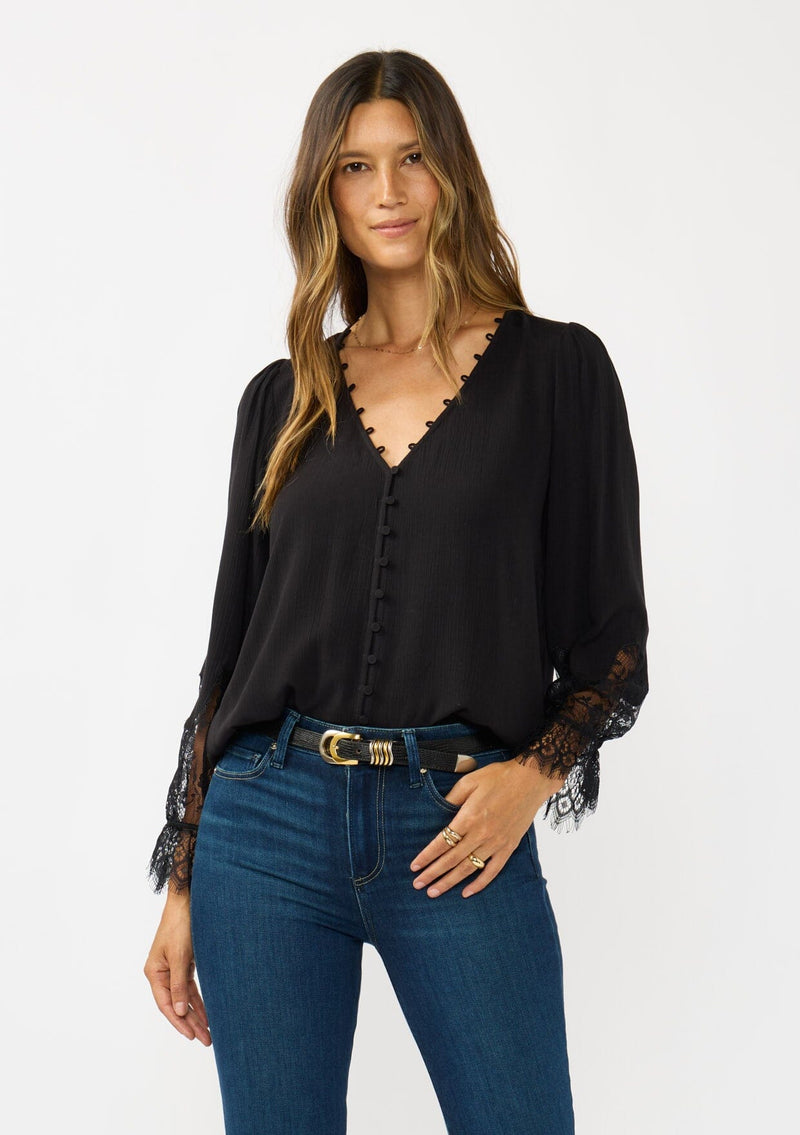 [Color: Black] A front facing image of a brunette model wearing a solid black blouse with lace accented sleeves, button front, and v-neckline. A classic and versatile boho blouse with an elevated touch. 