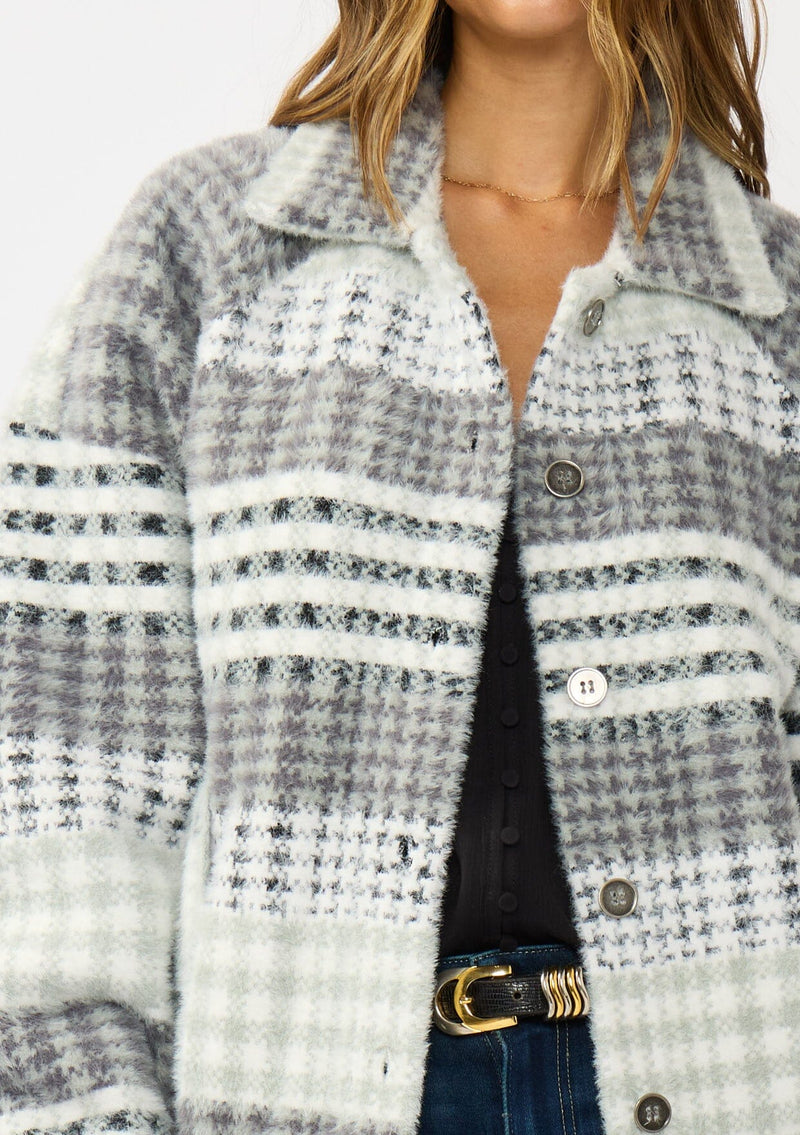 [Color: Grey/Cream] A detail image of a brunette model wearing a soft and fuzzy grey plaid bomber jacket with a collared neckline, button front, and side pockets. 