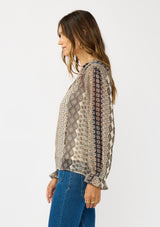 [Color: Natural/Navy] A side facing image of a brunette model wearing a beige striped floral pattern blouse with a relaxed boho silhouette, split v-neckline with ties, and long sleeves with ruffled elastic cuffs. 