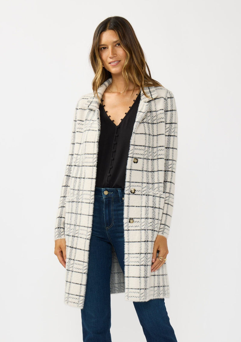 [Color: Cream/Black] A front facing image of a brunette model wearing a fuzzy statement sweater coat in a cream and black plaid print. With long sleeves, a notched collar, a snap button front, and side patch pockets. 