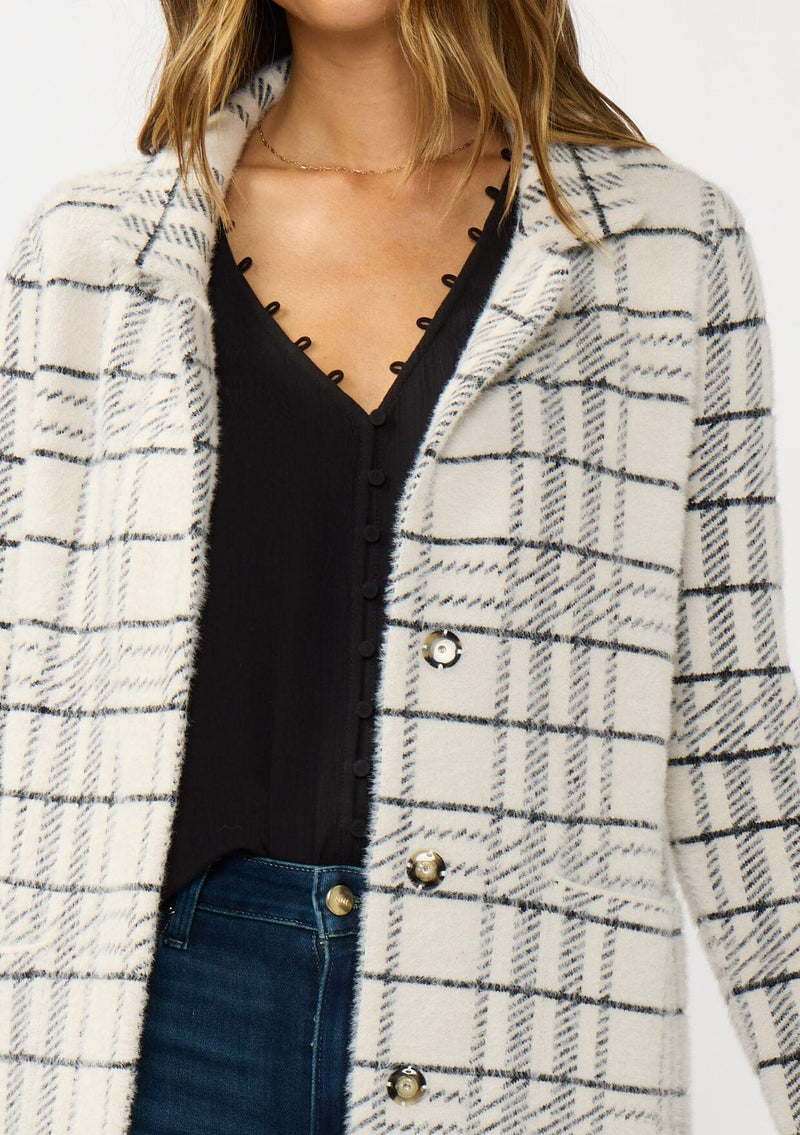 [Color: Cream/Black] A detail image of a brunette model wearing a fuzzy statement sweater coat in a cream and black plaid print. With long sleeves, a notched collar, a snap button front, and side patch pockets. 