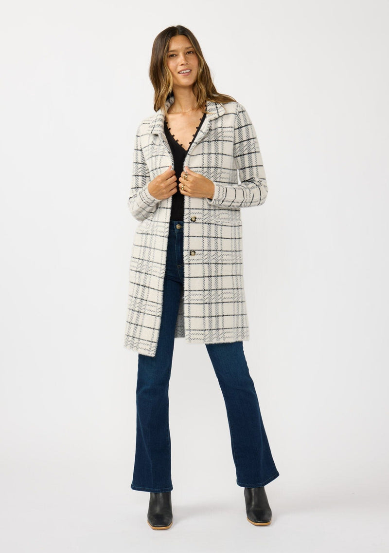 [Color: Cream/Black] A front facing image of a brunette model wearing a fuzzy statement sweater coat in a cream and black plaid print. With long sleeves, a notched collar, a snap button front, and side patch pockets. 