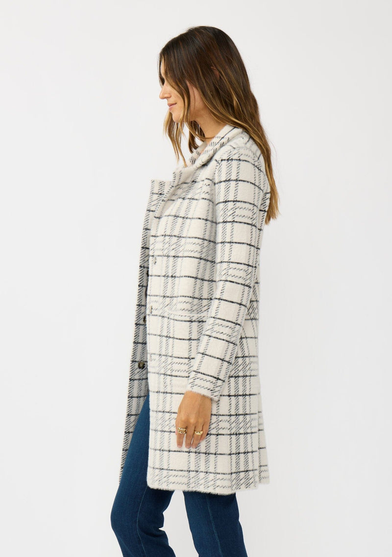 [Color: Cream/Black] A side facing image of a brunette model wearing a fuzzy statement sweater coat in a cream and black plaid print. With long sleeves, a notched collar, a snap button front, and side patch pockets. 
