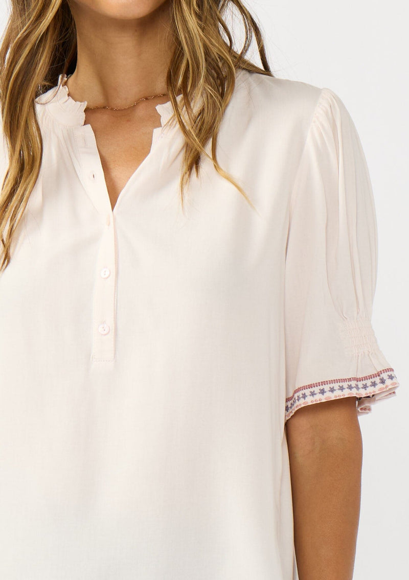 [Color: Pale Rose] A detail image of a brunette model wearing a light pink pink top with embroidered details on the puff sleeves, a button front, and a ruffled neck accent. 