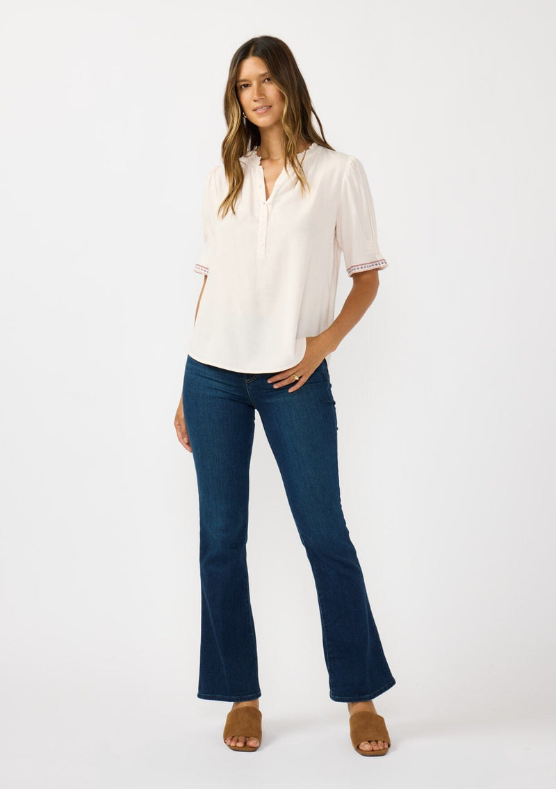 [Color: Pale Rose] A front facing image of a brunette model wearing a light pink pink top with embroidered details on the puff sleeves, a button front, and a ruffled neck accent. 