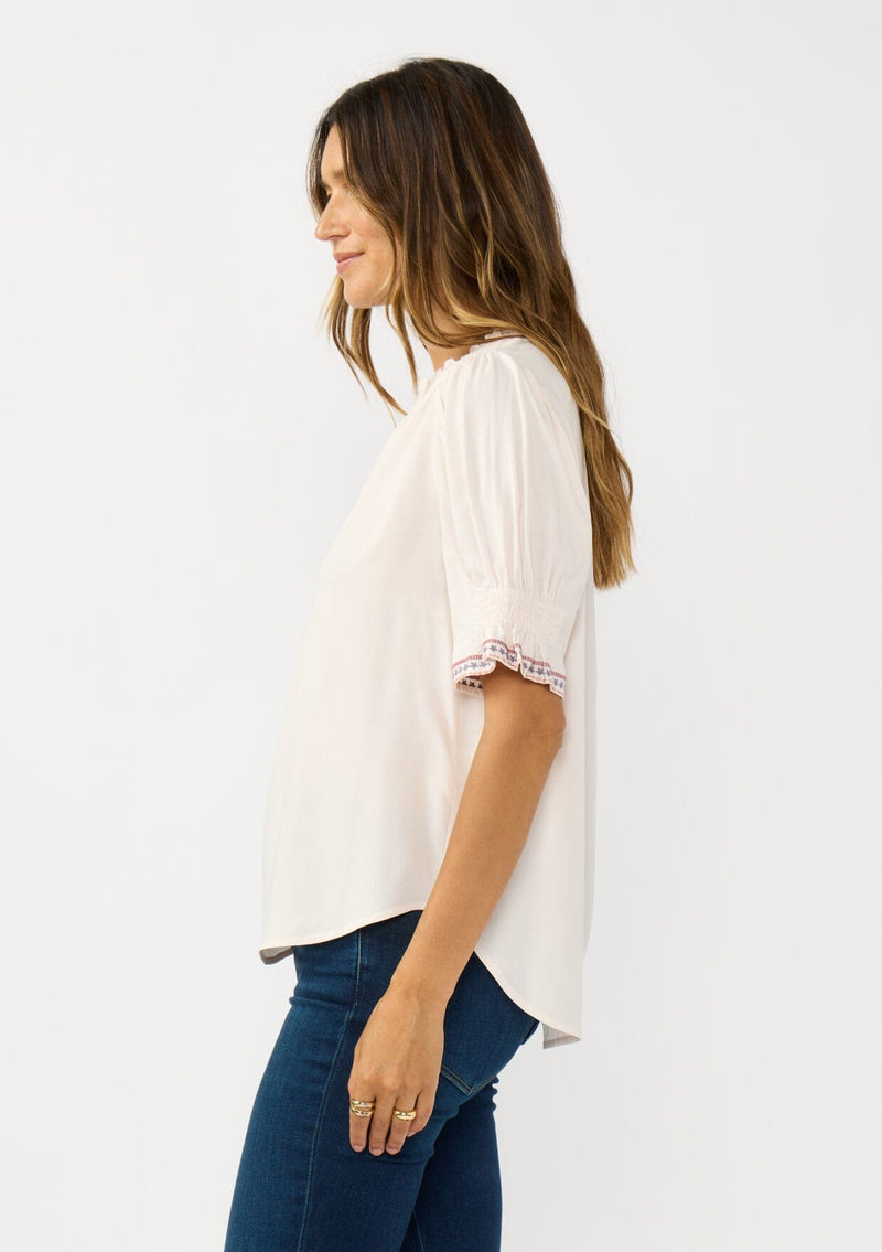 [Color: Pale Rose] A side facing image of a brunette model wearing a light pink pink top with embroidered details on the puff sleeves, a button front, and a ruffled neck accent. 