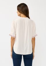 [Color: Pale Rose] A back facing image of a brunette model wearing a light pink pink top with embroidered details on the puff sleeves, a button front, and a ruffled neck accent. 