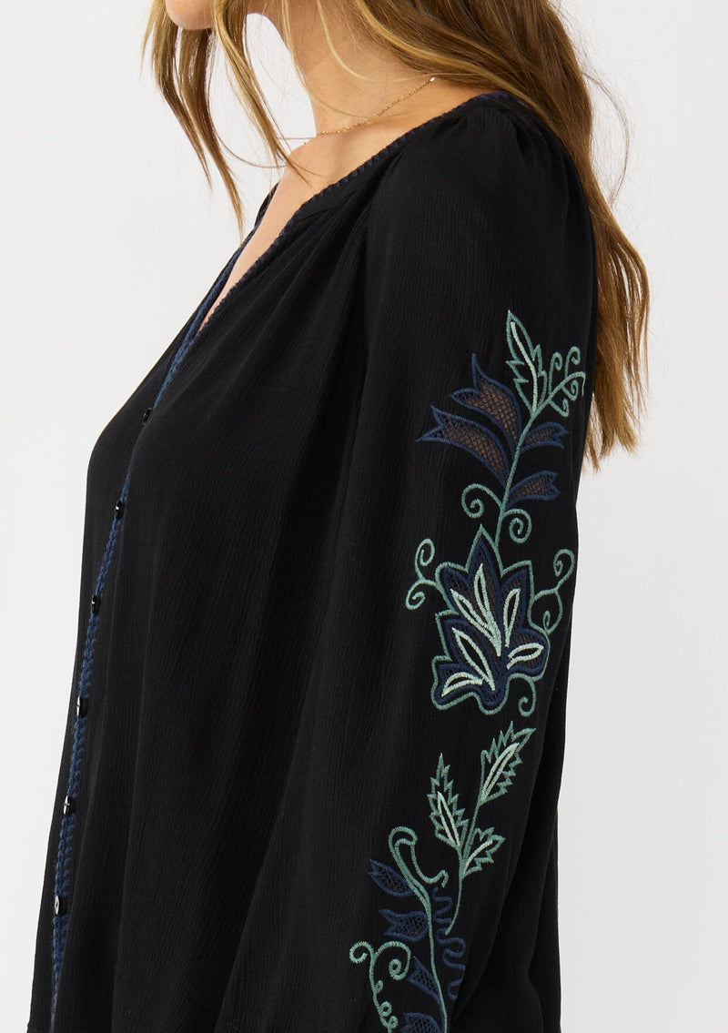 [Color: Black] A detail image of a brunette model wearing a flowy black blouse with colorful embroidered sleeve details, a button front, and blanket stitch detailing. Perfect for fall styling. 