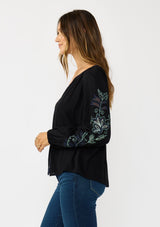 [Color: Black] A side facing image of a brunette model wearing a flowy black blouse with colorful embroidered sleeve details, a button front, and blanket stitch detailing. Perfect for fall styling. 