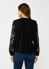 [Color: Black] A back facing image of a brunette model wearing a flowy black blouse with colorful embroidered sleeve details, a button front, and blanket stitch detailing. Perfect for fall styling. 