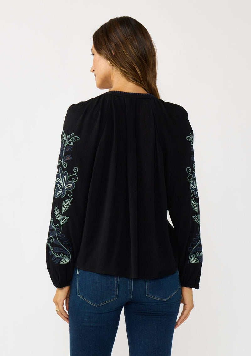 [Color: Black] A back facing image of a brunette model wearing a flowy black blouse with colorful embroidered sleeve details, a button front, and blanket stitch detailing. Perfect for fall styling. 