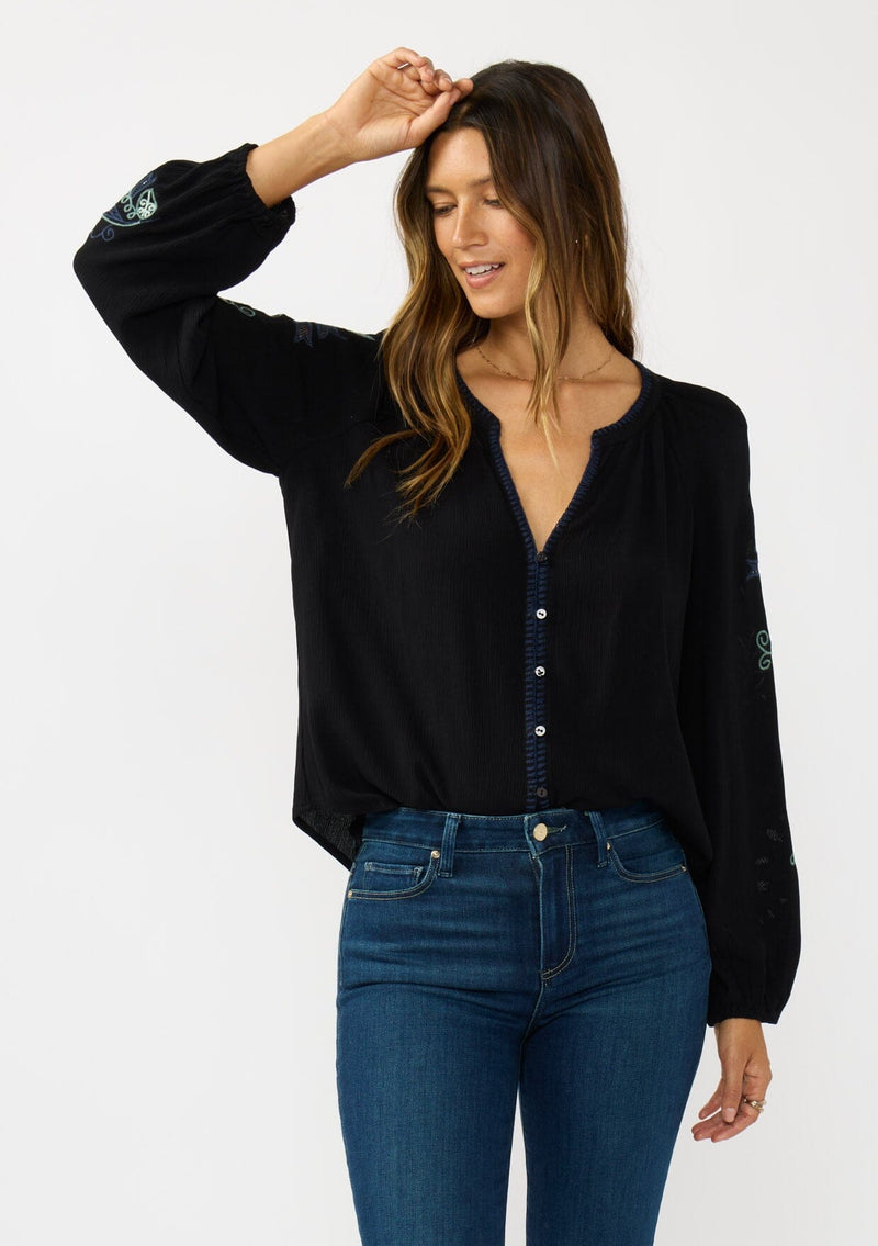 [Color: Black] A front facing image of a brunette model wearing a flowy black blouse with colorful embroidered sleeve details, a button front, and blanket stitch detailing. Perfect for fall styling. 
