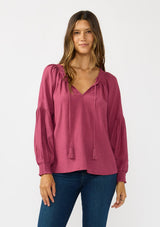 [Color: Berry] A front facing image of a brunette model wearing a pink boho blouse with metallic thread details. featuring a ruffled split v-neckline with tassel ties, long volume sleeves, and smocked cuffs. A fall boho blouse for casual days. 
