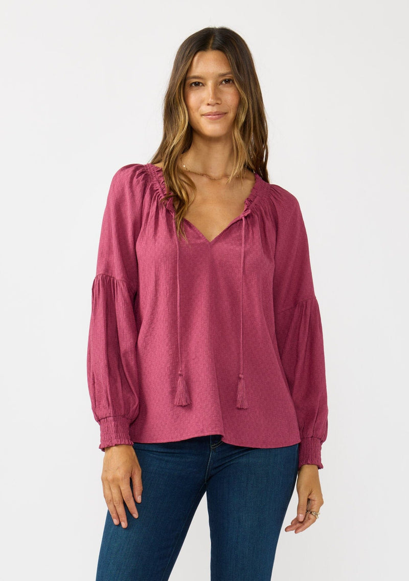 [Color: Berry] A front facing image of a brunette model wearing a pink boho blouse with metallic thread details. featuring a ruffled split v-neckline with tassel ties, long volume sleeves, and smocked cuffs. A fall boho blouse for casual days. 
