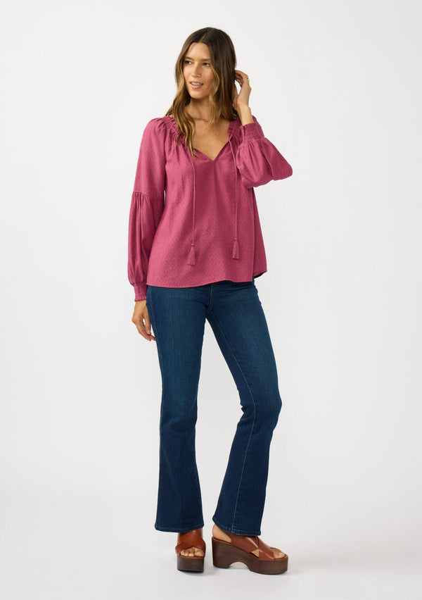 [Color: Berry] A front facing image of a brunette model wearing a pink boho blouse with metallic thread details. featuring a ruffled split v-neckline with tassel ties, long volume sleeves, and smocked cuffs. A fall boho blouse for casual days. 