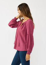 [Color: Berry] A side facing image of a brunette model wearing a pink boho blouse with metallic thread details. featuring a ruffled split v-neckline with tassel ties, long volume sleeves, and smocked cuffs. A fall boho blouse for casual days. 