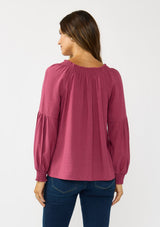 [Color: Berry] A back facing image of a brunette model wearing a pink boho blouse with metallic thread details. featuring a ruffled split v-neckline with tassel ties, long volume sleeves, and smocked cuffs. A fall boho blouse for casual days. 