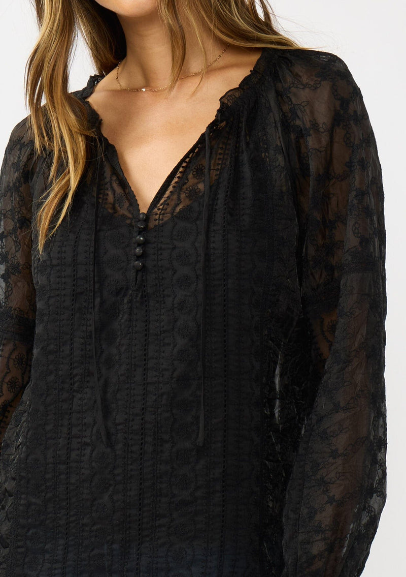 [Color: Black] A detail image of a brunette model wearing a sheer crinkled chiffon blouse with all over embroidery. Featuring a split v-neckline with tie detail and a button front closure.