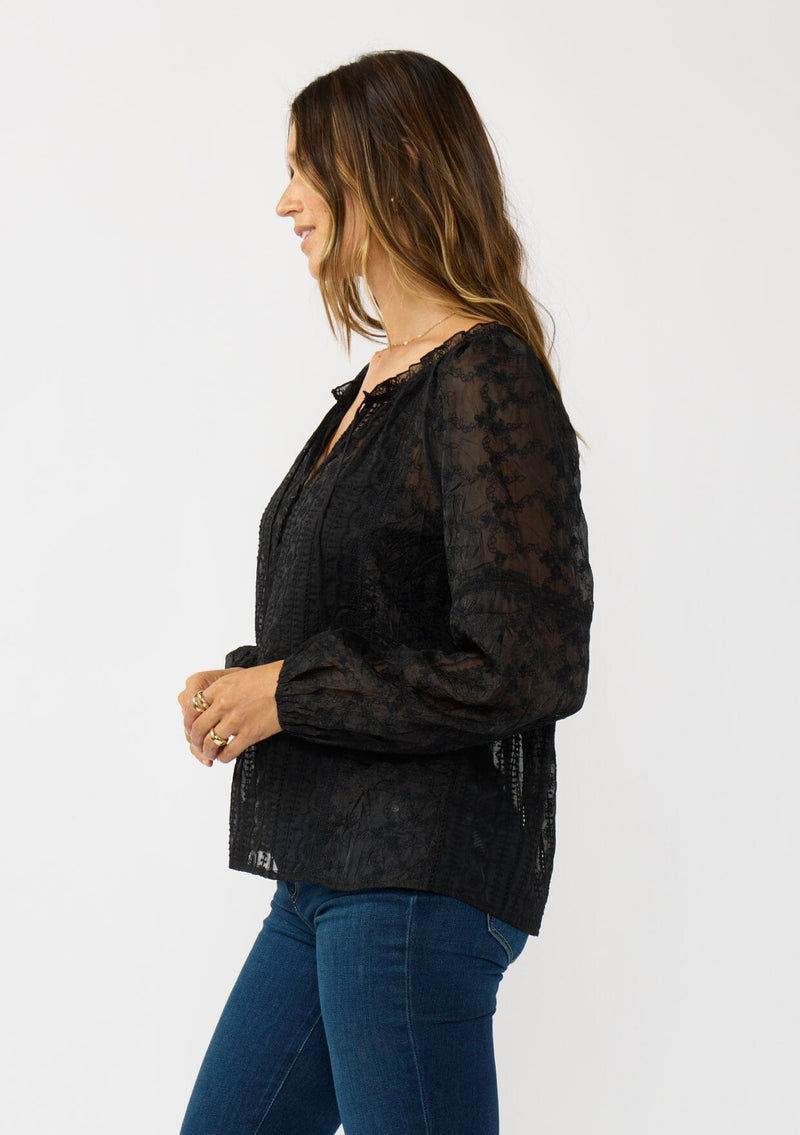 [Color: Black] A side facing image of a brunette model wearing a sheer crinkled chiffon blouse with all over embroidery. Featuring a split v-neckline with tie detail and a button front closure. 